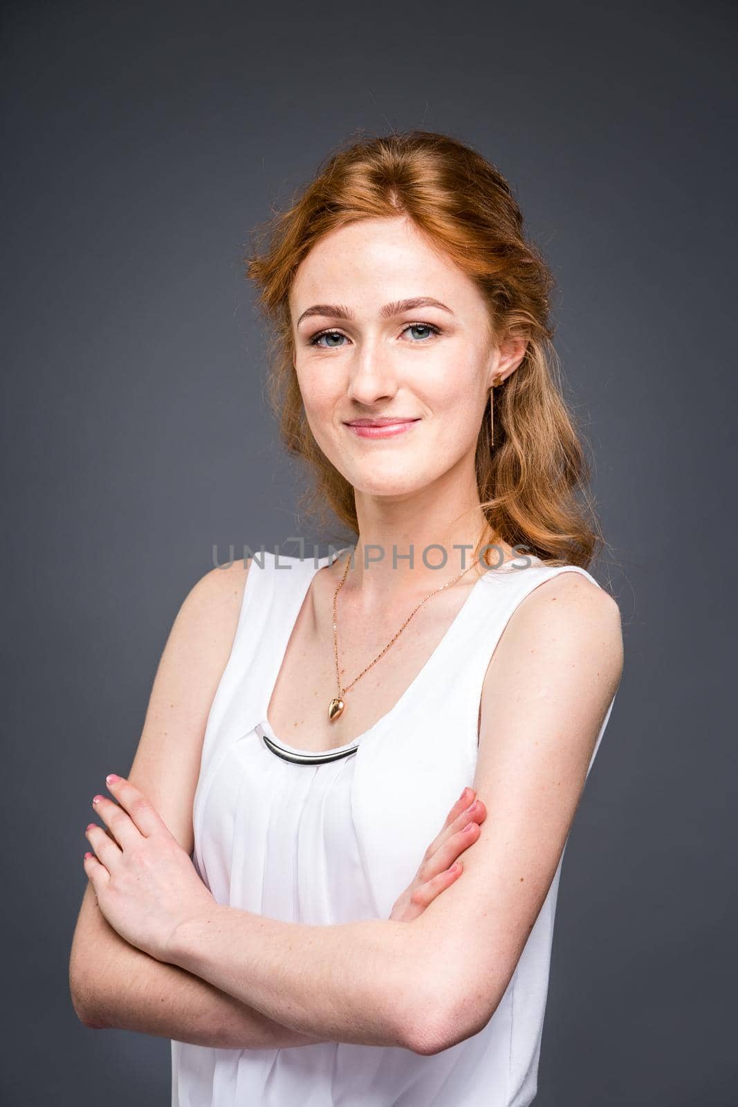 portrait of a young red-haired beautiful girl in the studio on a gray isolated background. A woman is standing with her arms folded and smiling in a white shirt with a short sleeve. Business concept