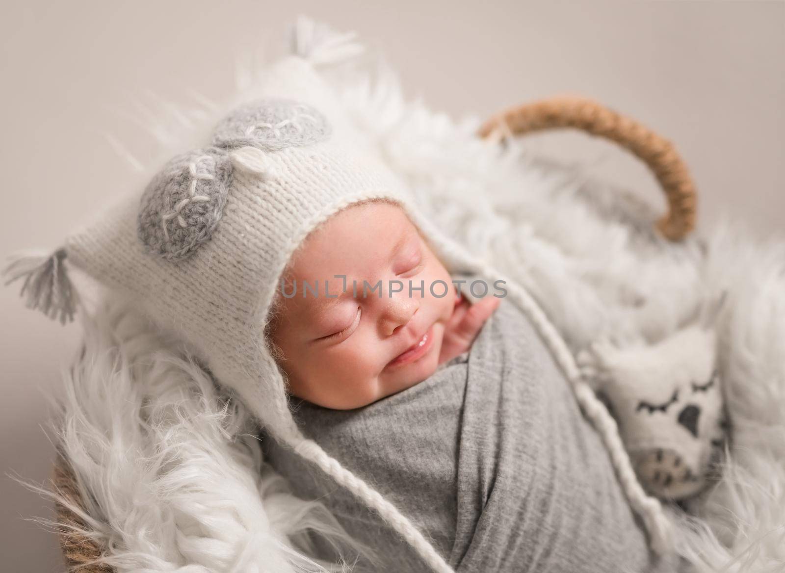 Little cute baby in white knitted beanie covered with gray coverlet sweetly sleeping on white soft blanket with toy owl in the basket