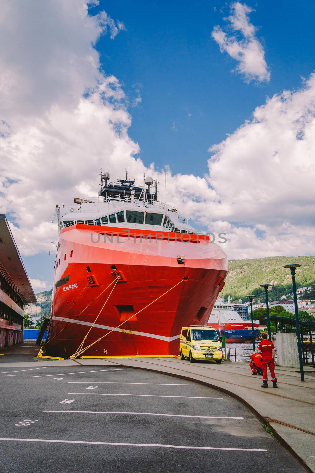 Two Norwegian paramedics in red uniforms are resting near an ambulance parked in a port near a large ship. Theme healthcare and medicine in Norway, Bergen July 28, 2019 by Tomashevska