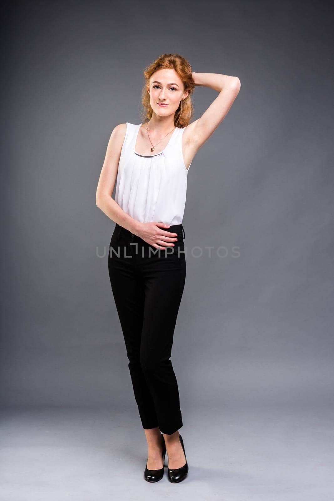 beautiful young Caucasian woman with long red hair in high heels, black trousers and a white shirt stands in full growth on a gray isolated background. by Tomashevska