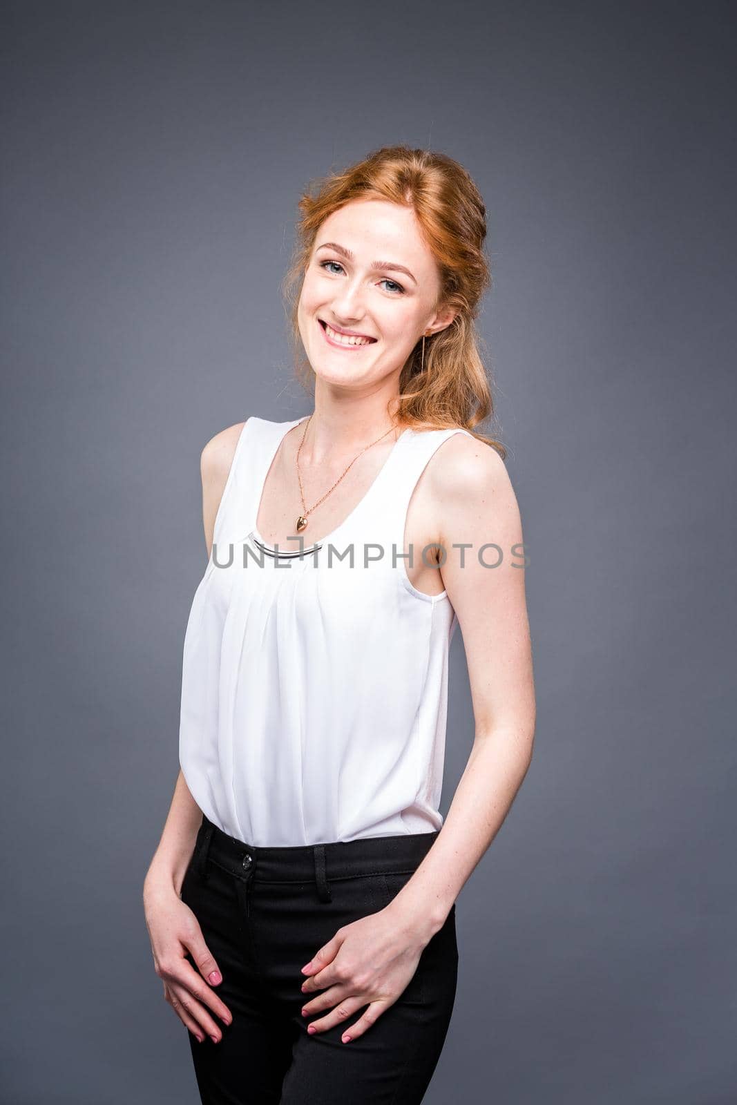 portrait of a young redhaired beautiful girl in the studio on a gray isolated background. A woman is standing with her arms folded and smiling in a white shirt with a short sleeve. Business concept by Tomashevska