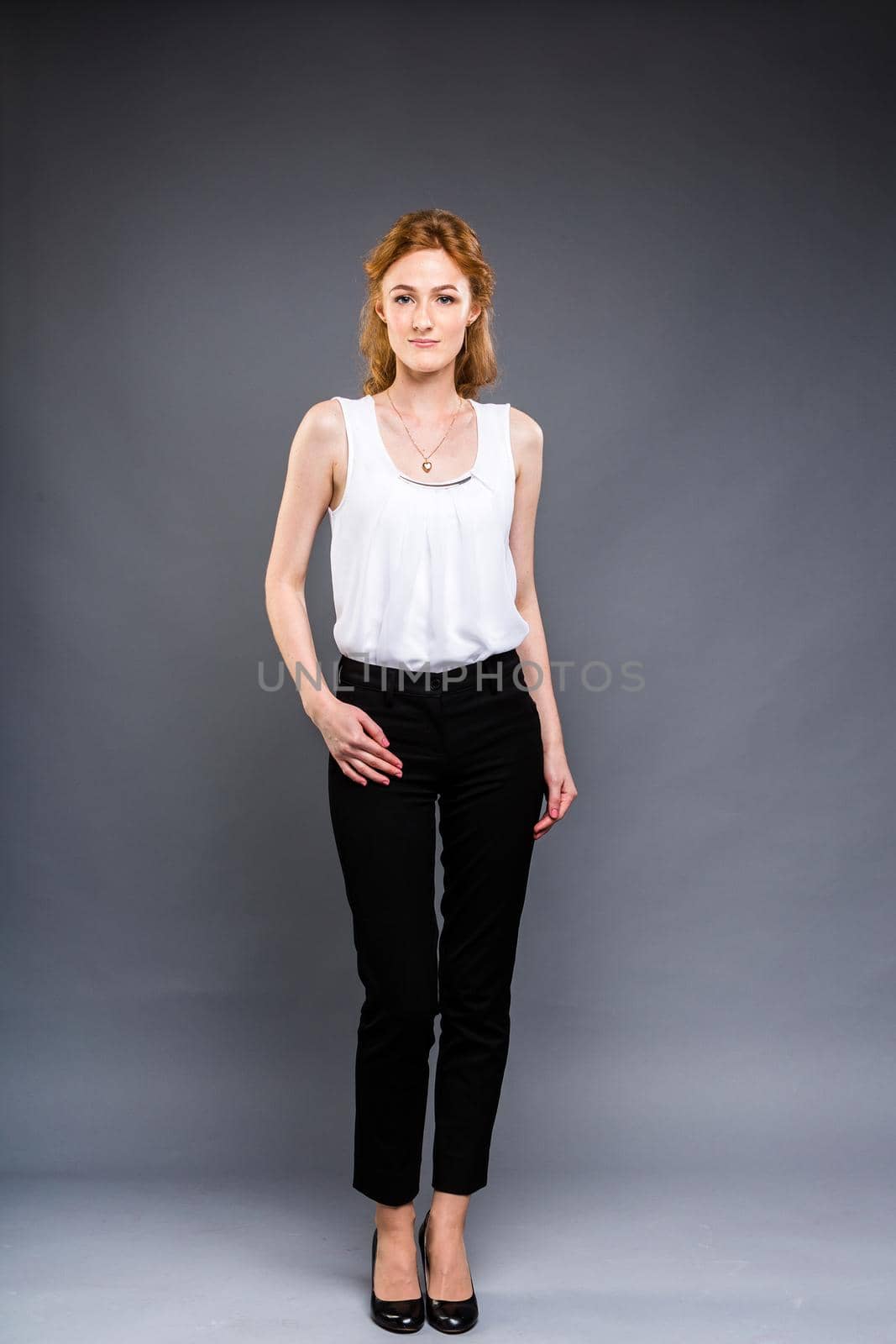 beautiful young Caucasian woman with long red hair in high heels, black trousers and a white shirt stands in full growth on a gray isolated background. Business concept