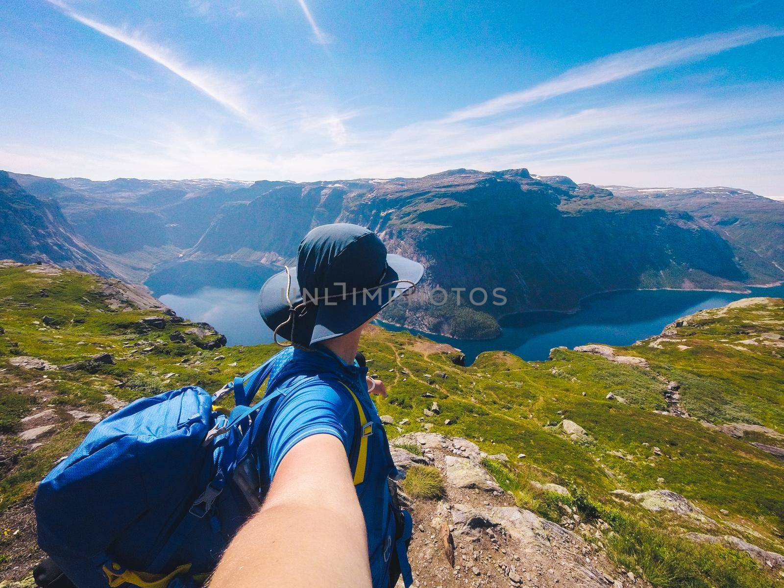 Ringedalsvatnet Lake Near Trolltunga. Blue Lake In Norway. Woman tourist in hat and backpack stands back and holds camera in hand takes selfie photo on background mountains and lake nature.