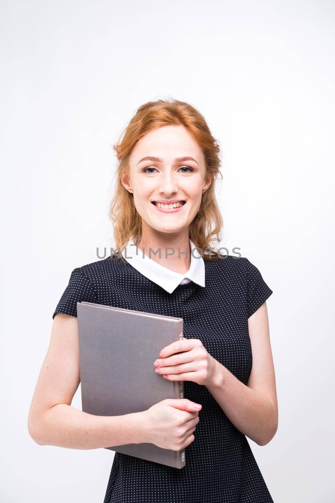 Beautiful girl with red hair and gray book in hands dressed in black dress on white isolated background by Tomashevska