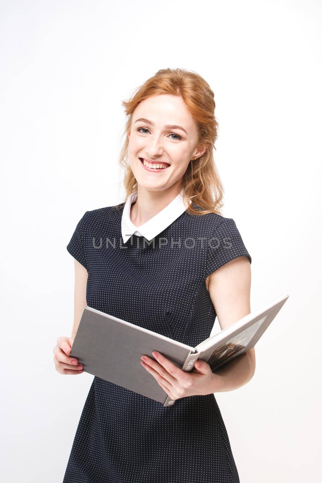 Beautiful girl with red hair and gray book in hands dressed in black dress on white isolated background.
