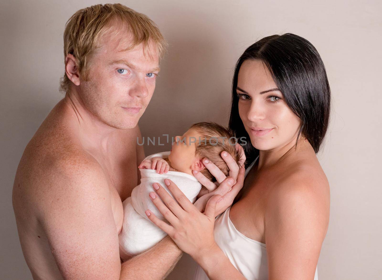 Father and mother holding little sleeping baby together covered in white coverlet on the light background
