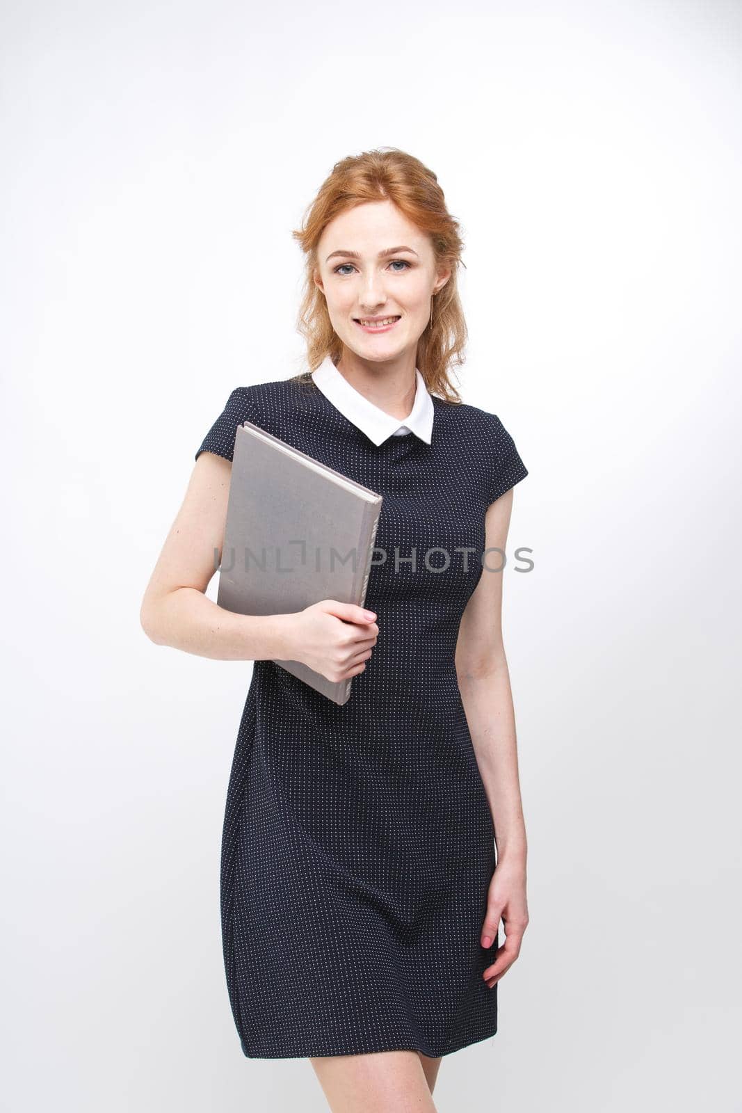Beautiful girl with red hair and gray book in hands dressed in black dress on white isolated background by Tomashevska