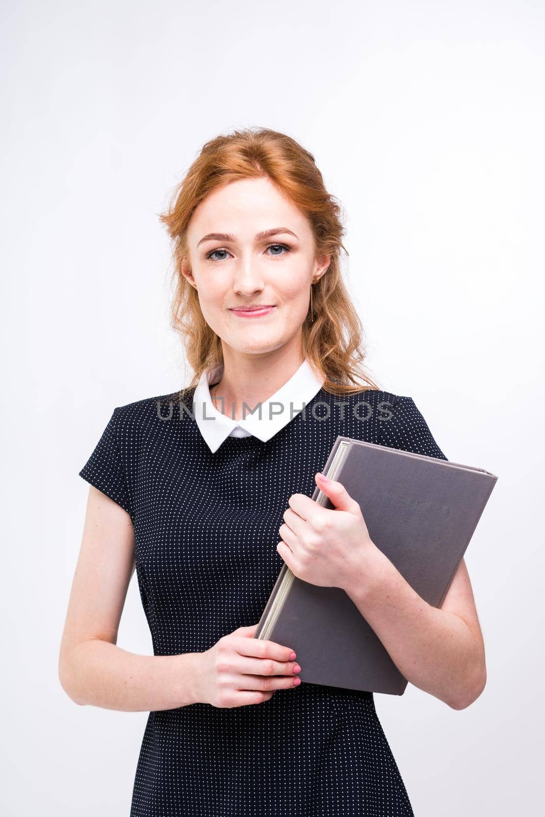 Beautiful girl with red hair and gray book in hands dressed in black dress on white isolated background by Tomashevska