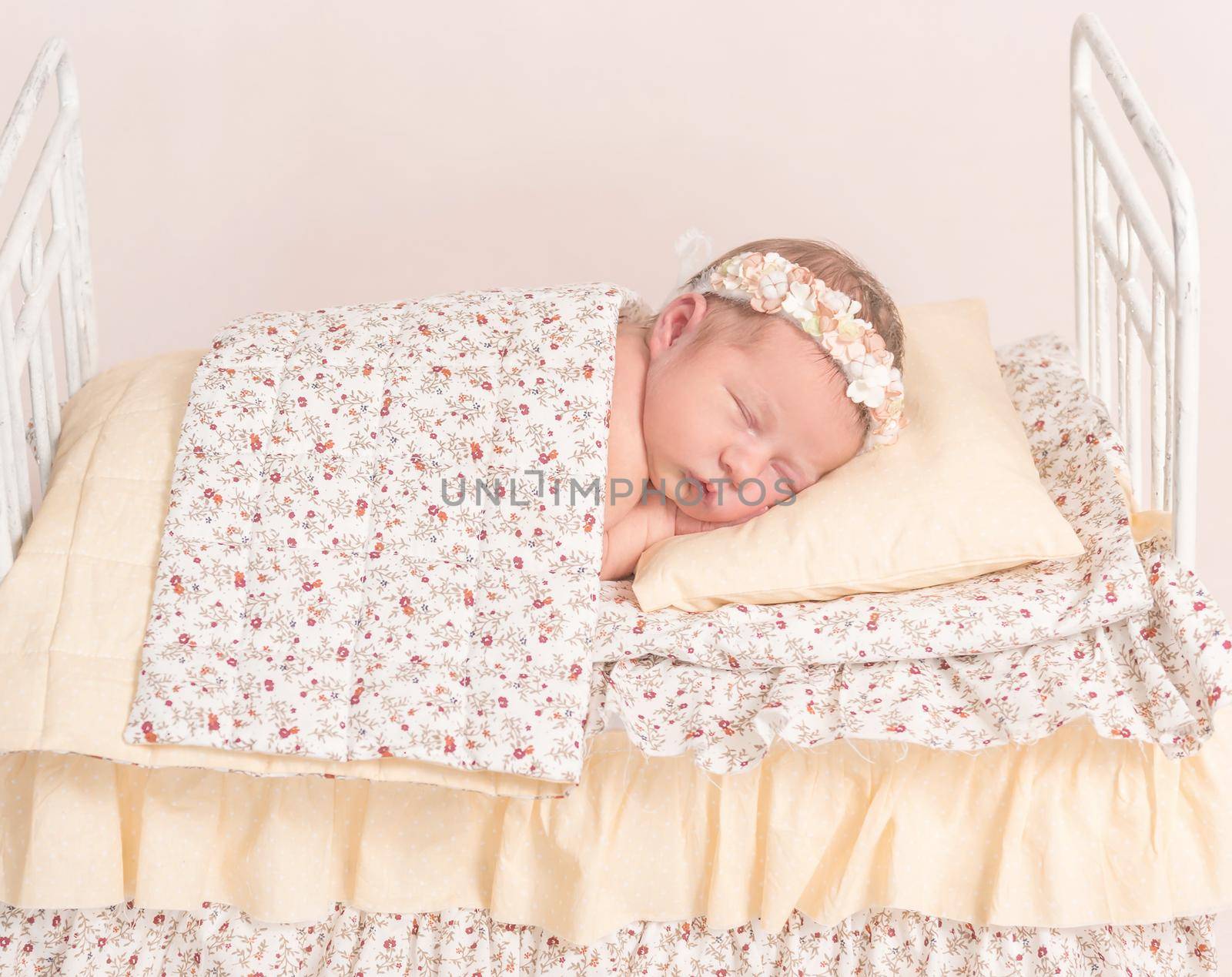 Infant in hairband napping under the sheets of lovely kids' bed with yellowish sheets