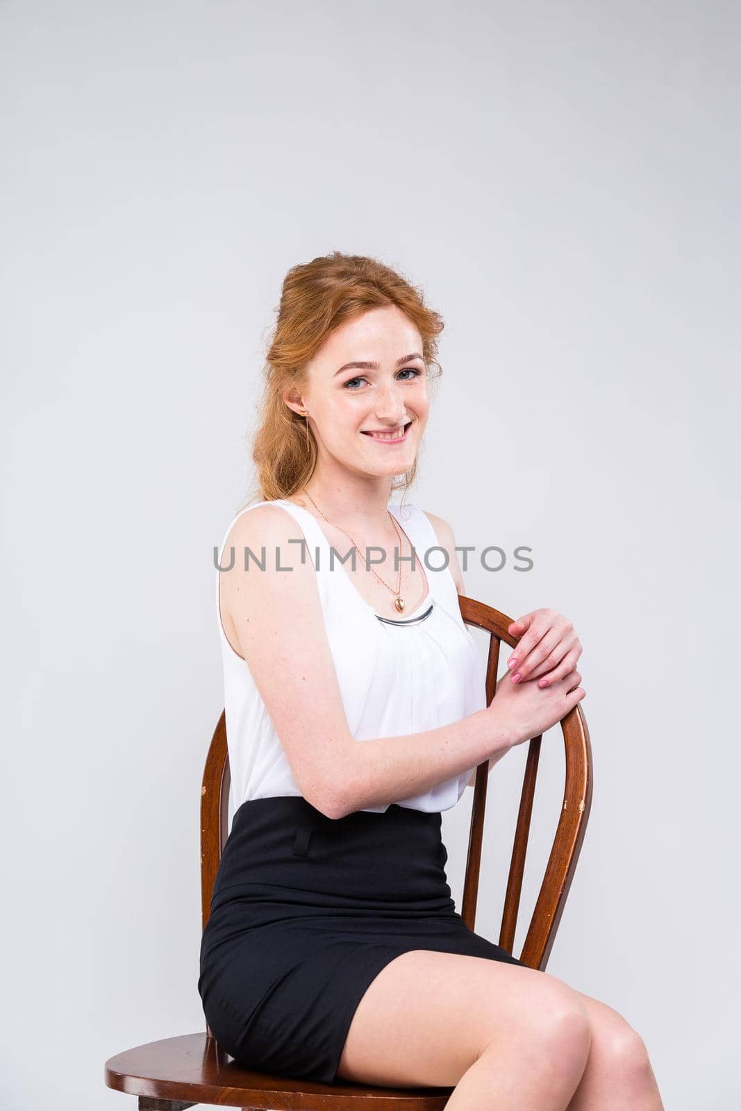 Young beautiful woman with long red, curly hair sitting on a wooden chair on a white background in the studio. Dressed in a white blouse with a short sleeve and a black short skirt and high heels by Tomashevska