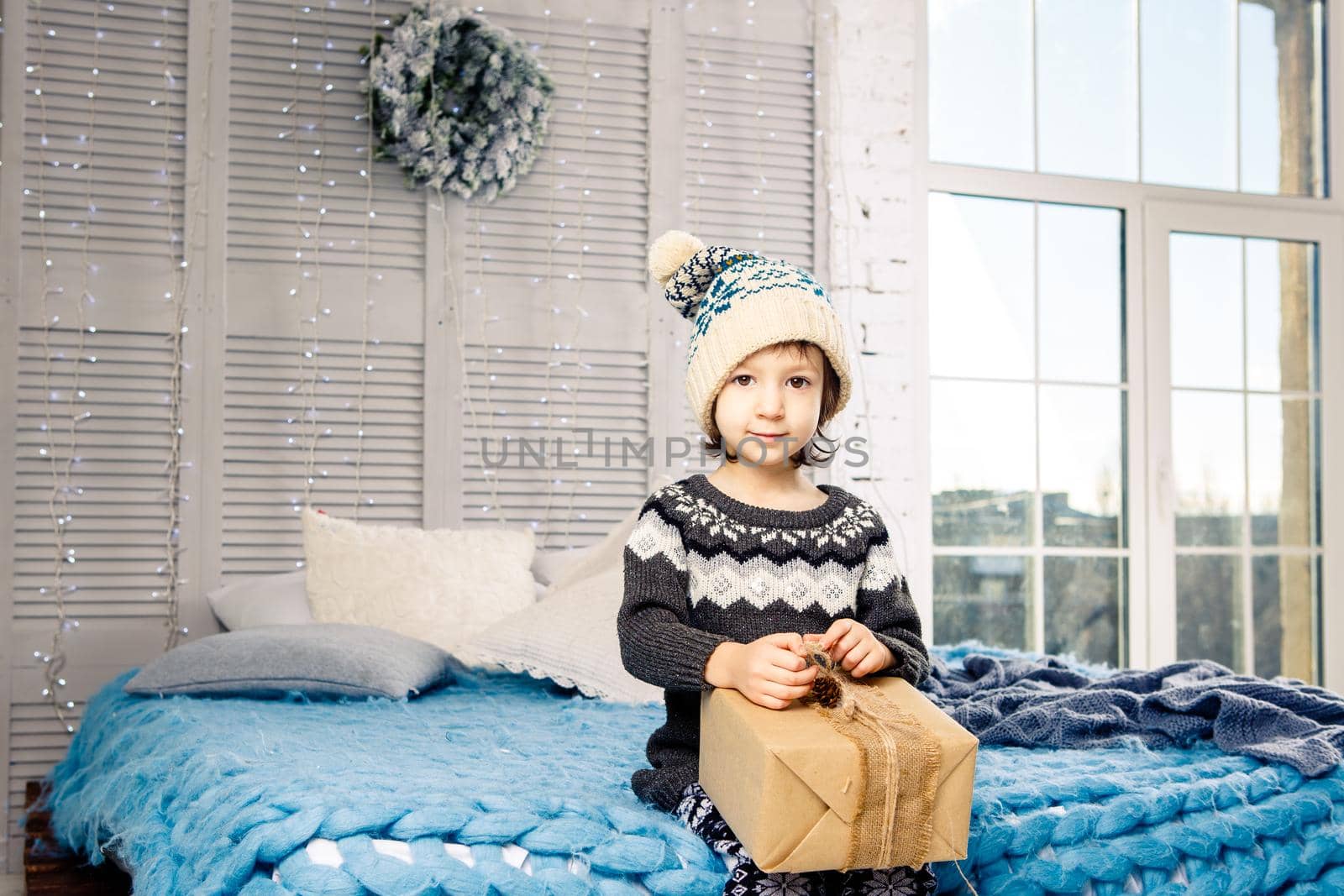 little girl the child sitting in pajamas and hat on the bed with garland of light bulbs with gifts boxes wrapped in a non-colored paper decorated with cones on blue knitted coverlet.Christmas concept.