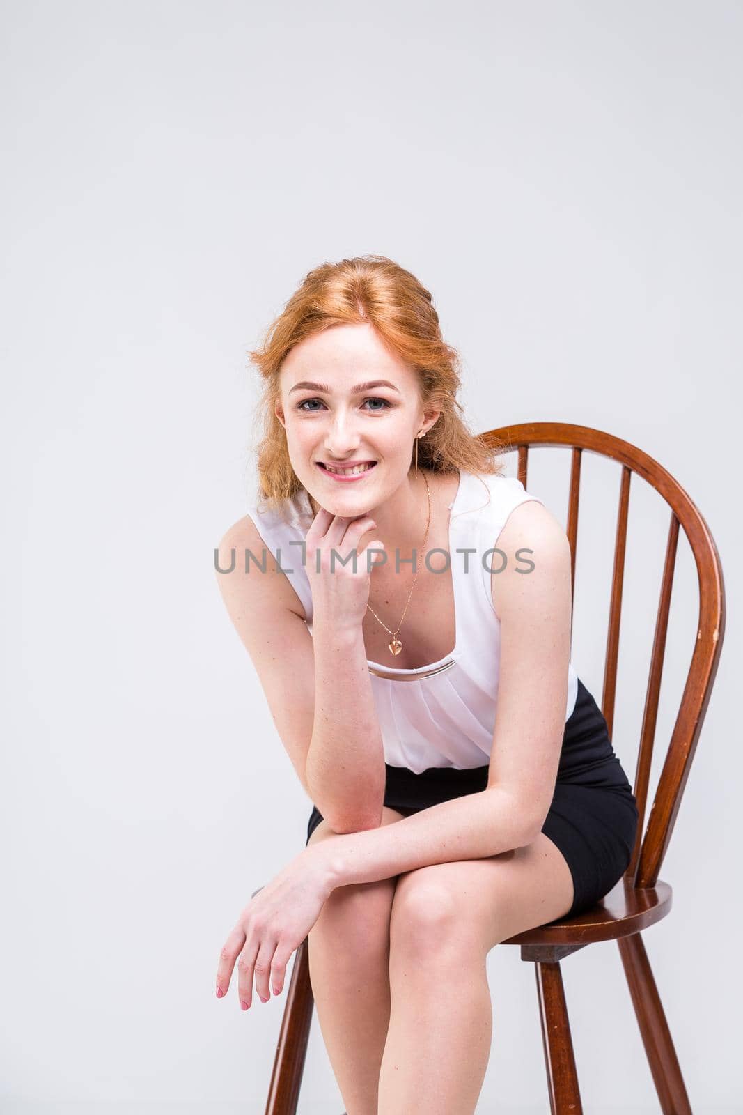 Portrait of beautiful business woman with long red, curly hair sitting on wooden chair on white background in studio. Dressed in a white blouse with a short sleeve and black short skirt and high heels