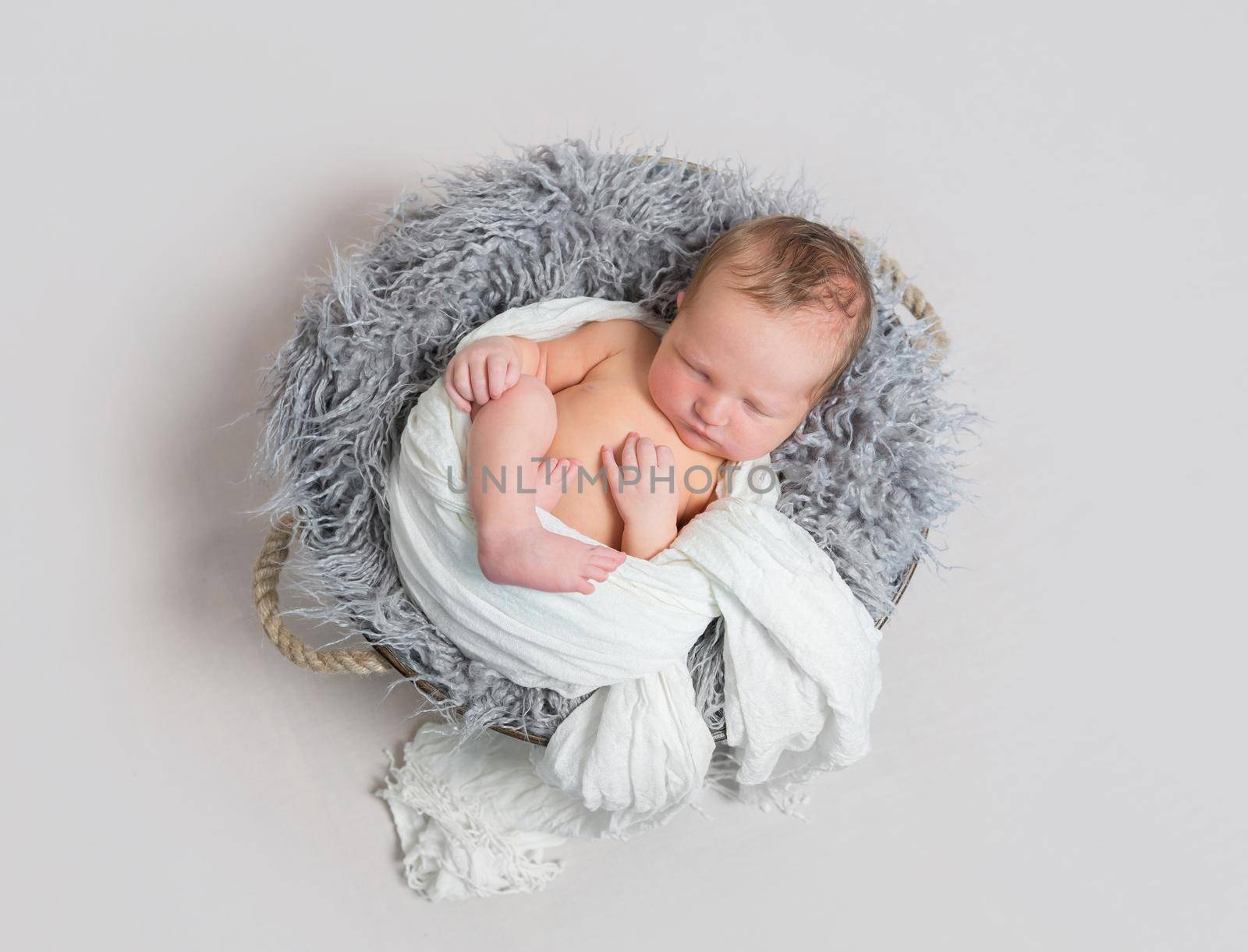 Lovely baby sleeping half wrapped up with white scarf, in a basket on gray pillow