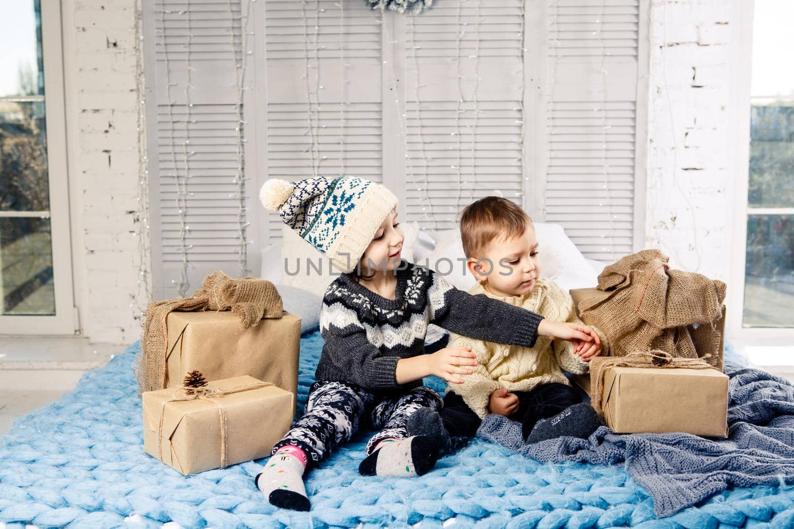 Theme Christmas morning. Two child Caucasian boy and girl brother and sister are sitting on the bed in an embrace with a smile and joy are opening New Year's gifts in boxes on a sunny day.