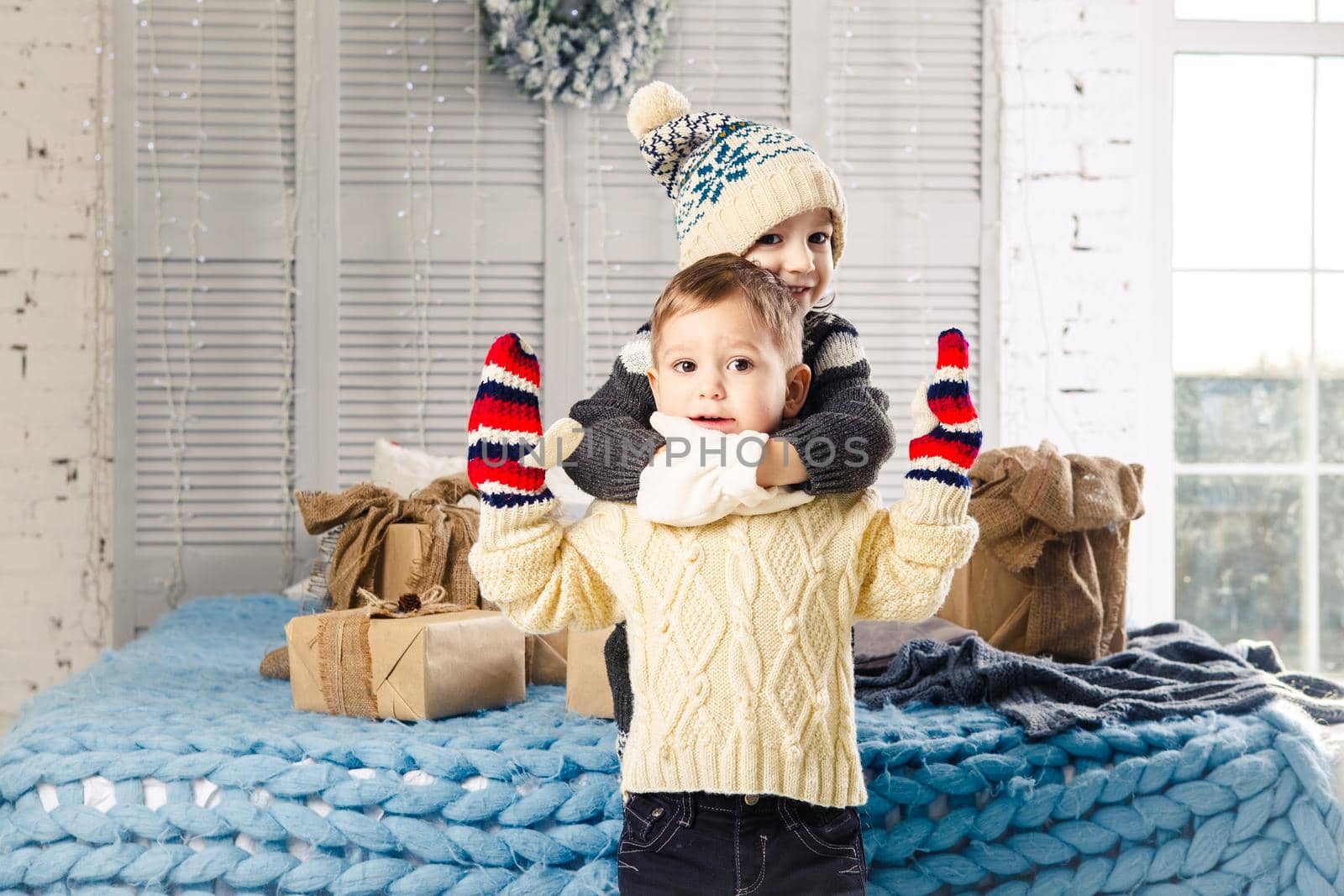 Christmas concept.Children brother and sister in embrace of the house in the bedroom near bed with boxes,gifts on back of the decor and windows on sunny day.Wear a warm woolen garment cap and mittens by Tomashevska