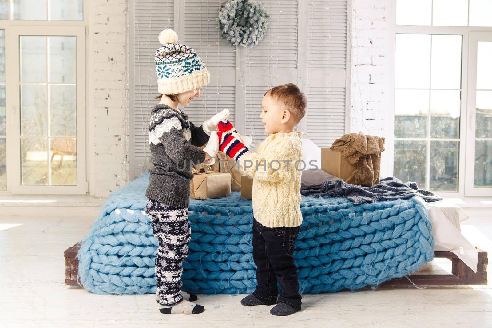 Children brother and sister standing and playing at home in the bedroom near bed with boxes, gifts on the background of Christmas decor a sunny day.Wear a warm knitted woolen garment cap and mittens by Tomashevska