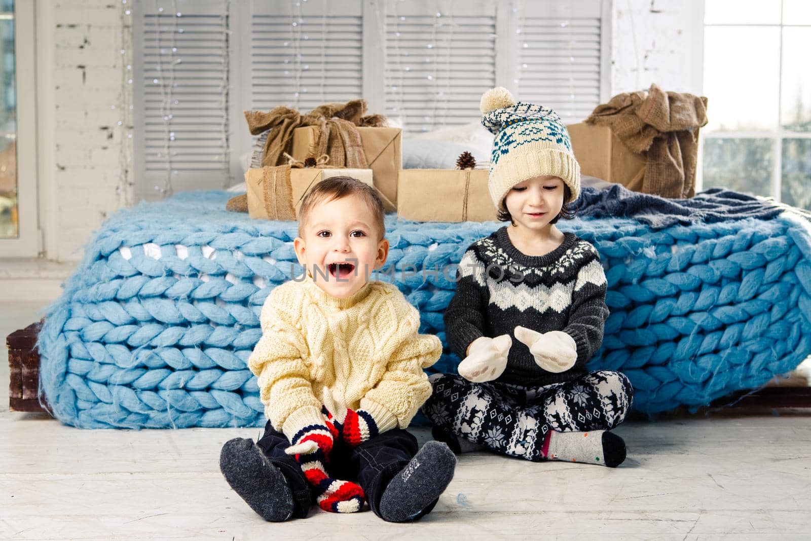 Children brother and sister sit on the floor in bedroom near the bed with gifts on the background of Christmas decor on a sunny day. Dressed in a warm knitted woolen garment cap and colored mittens.