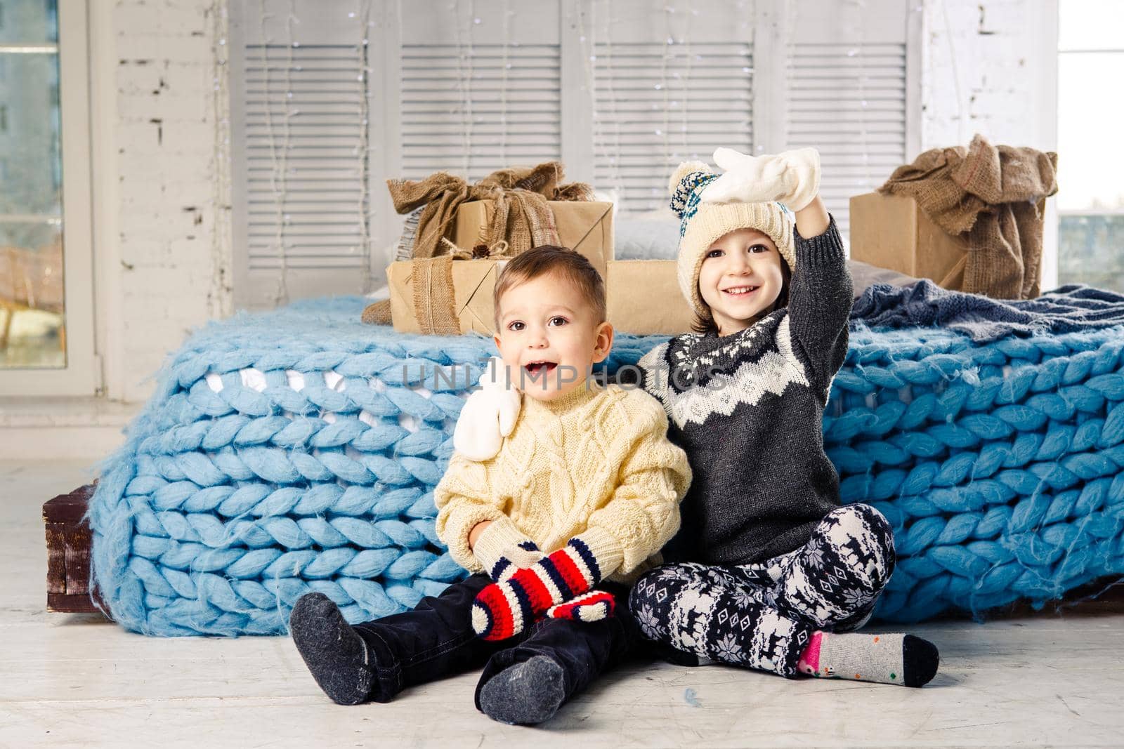 Children brother and sister sit on the floor in bedroom near the bed with gifts on the background of Christmas decor on a sunny day. Dressed in a warm knitted woolen garment cap and colored mittens by Tomashevska