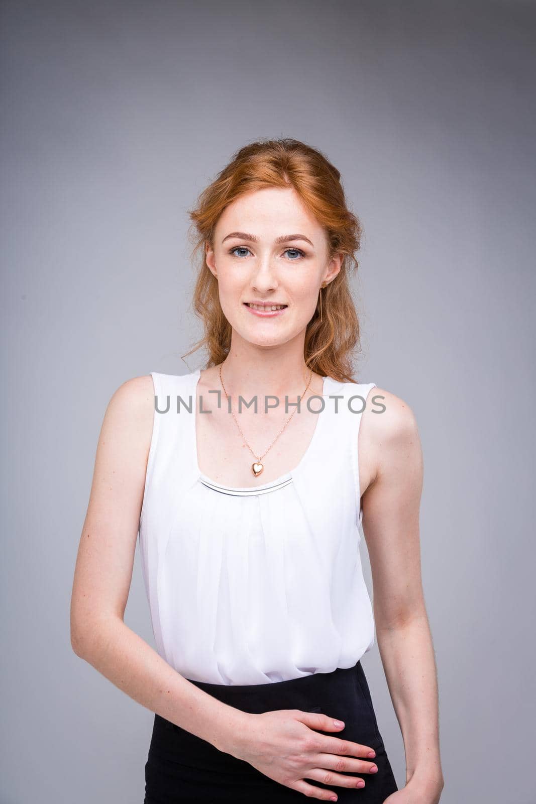portrait vertical up to the waist young, beautiful business woman, student with lred, curly hair and freckles on gray background. SHe dressed in a white blouse with short sleeves about open shoulders by Tomashevska