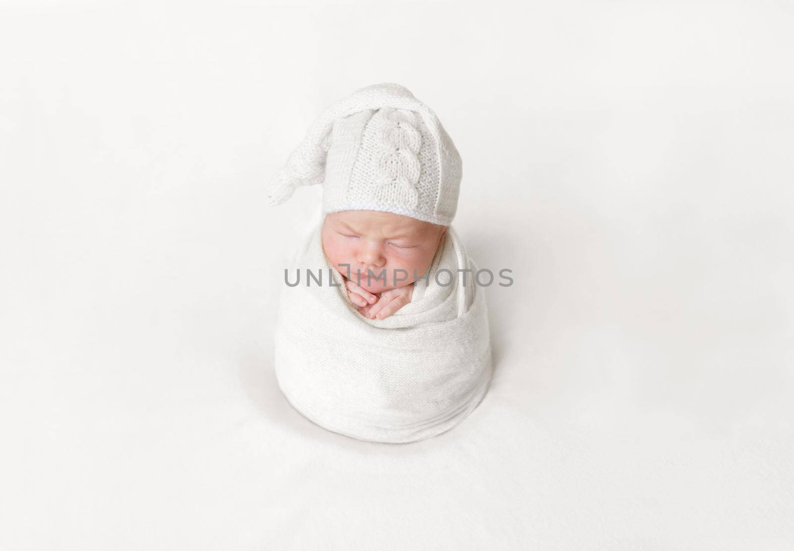 Lovely baby sleeping wrapped up with a blanket into a sitting position, white hat