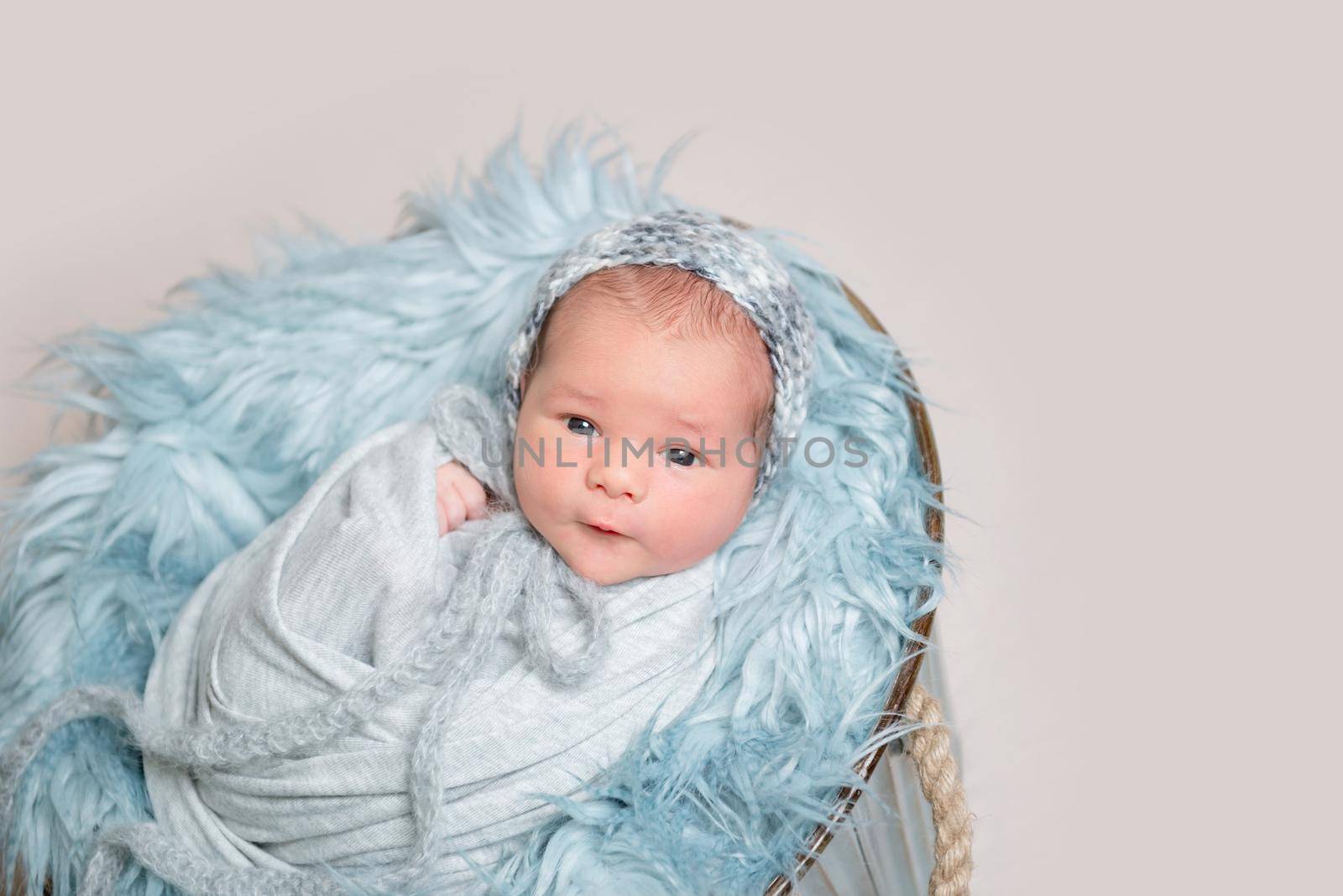 Newborn baby lying on basket covered with blue fluffy rug and curiously looking at camera, top view.