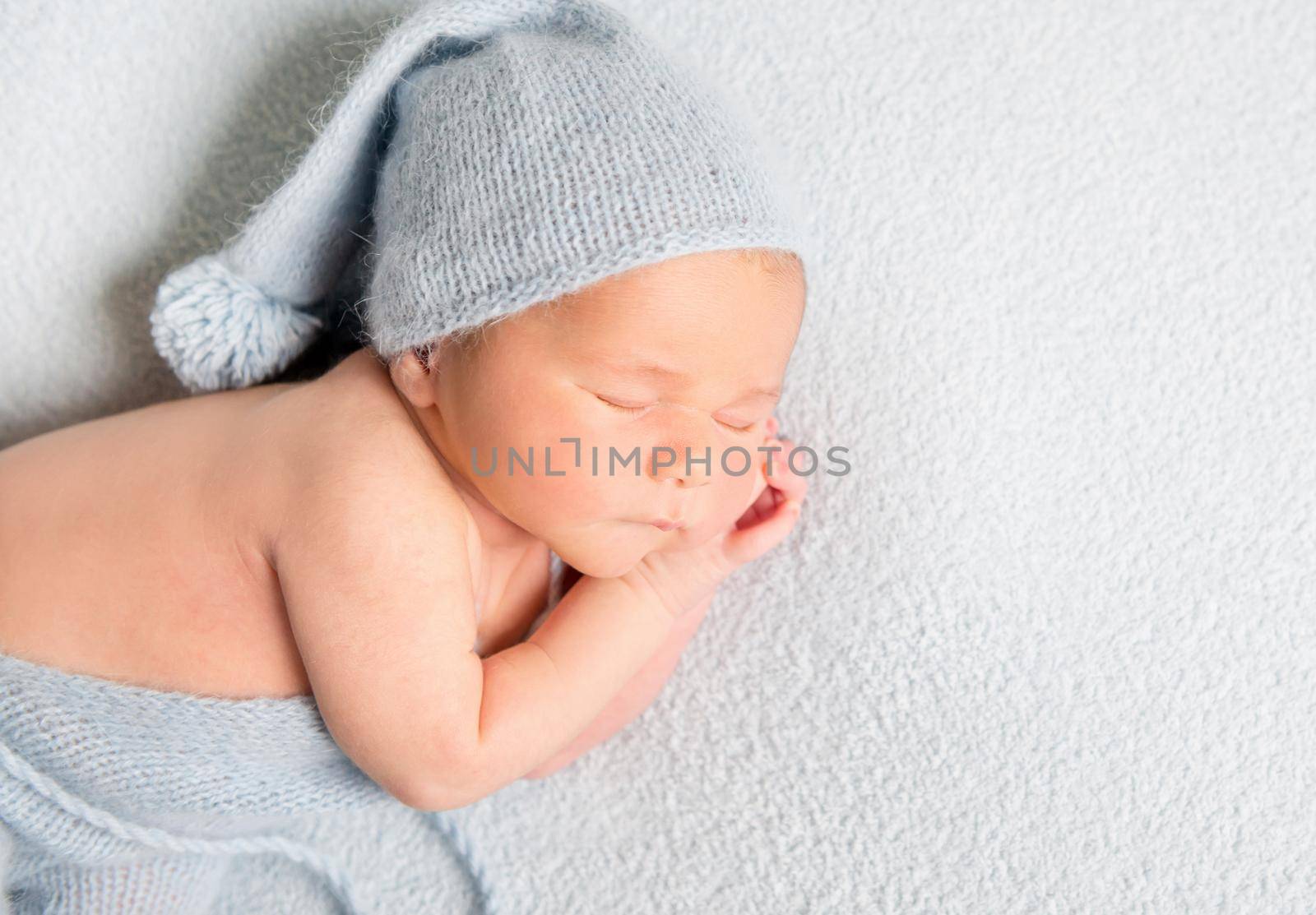 Sleeping baby boy in blue bonnet curled up on white blanket.