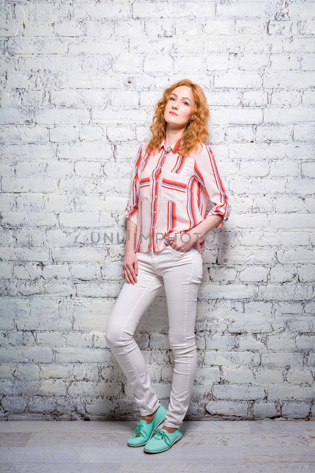 Full-length portrait A young beautiful Caucasian woman student stands near a white brick wall. Girl with red long curly hair with freckles on her face in bright summer clothes by Tomashevska
