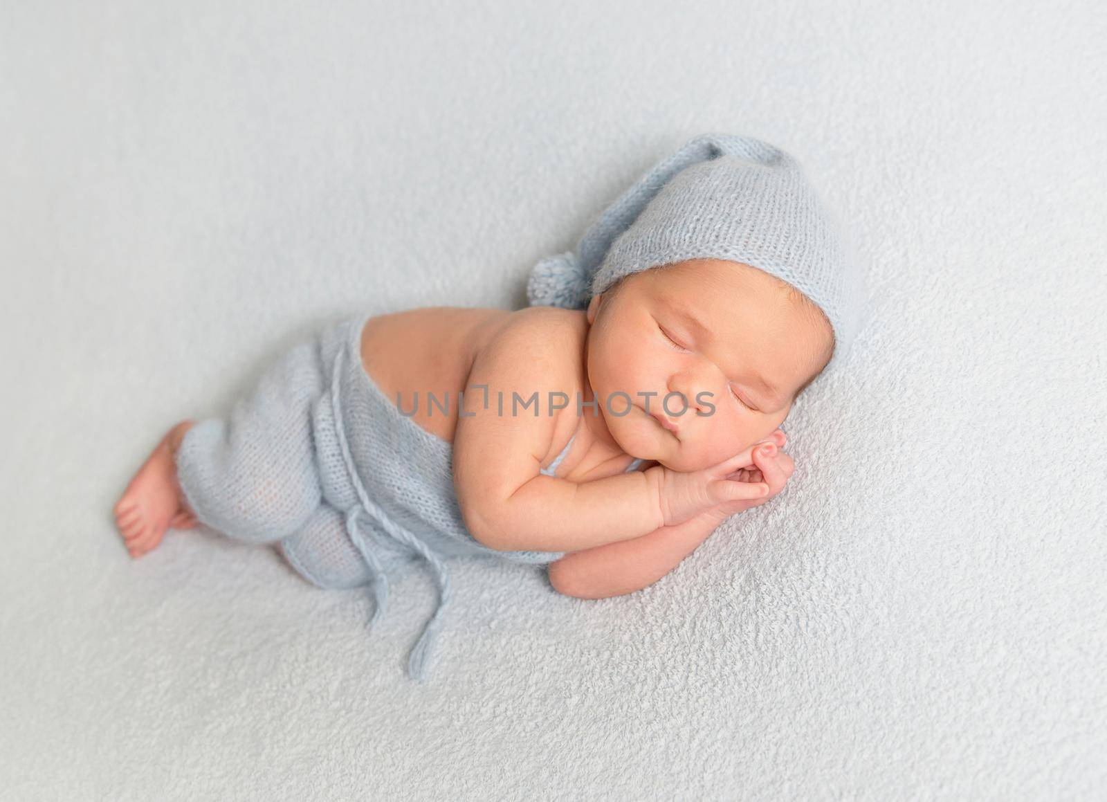 Sleeping baby boy in blue bonnet curled up on white blanket.