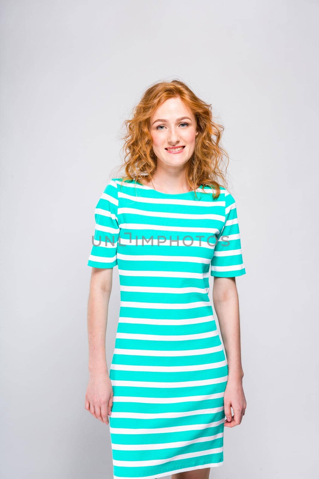 A lap portrait of a young, beautiful woman with red curly hair in a summer dress with stripes of blue in the studio on a gray background. Theme of summer vacation, tourism and summer clothes.