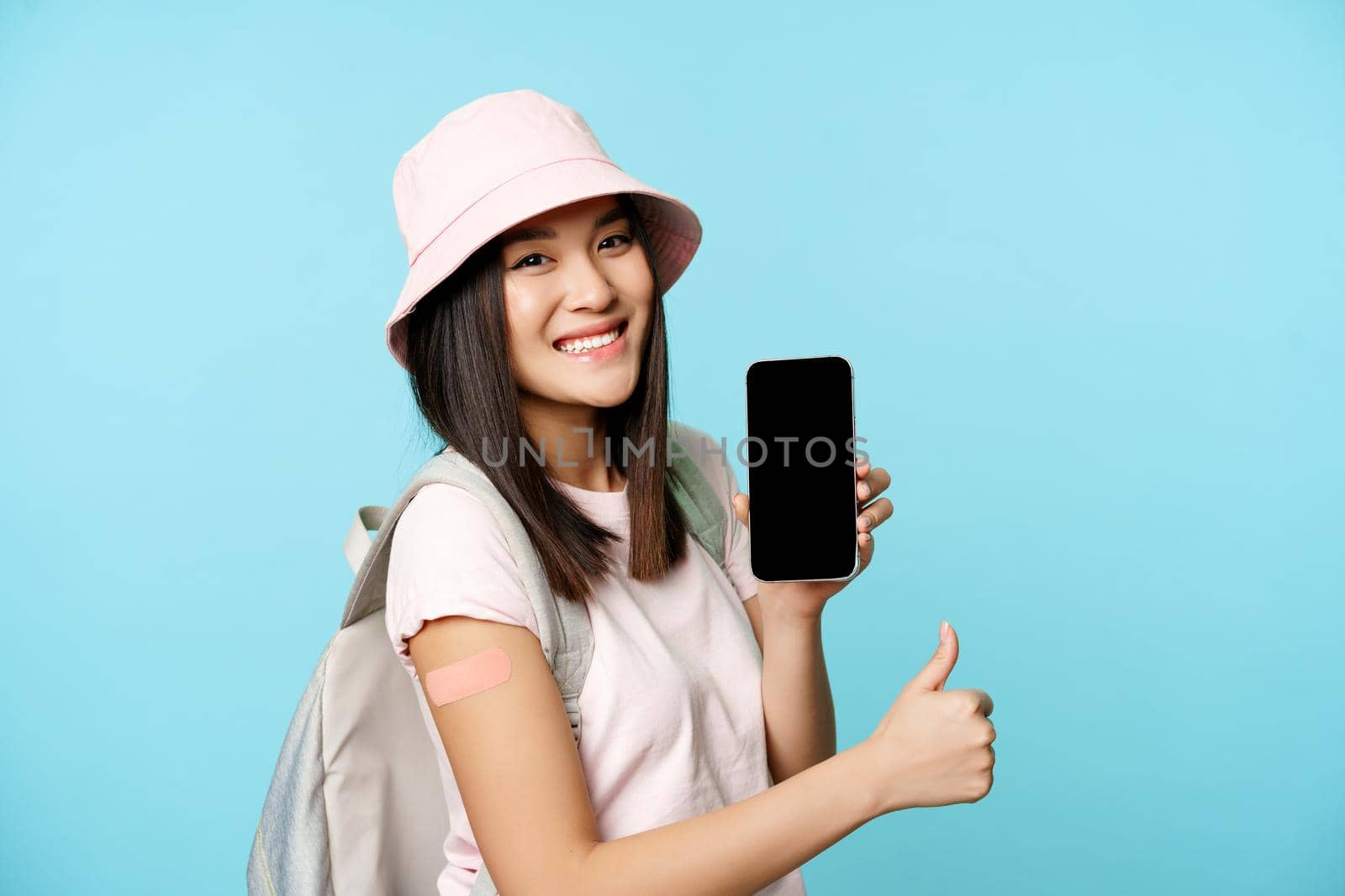 Smiling asian tourist shows arm with patch from covid-19 vaccination, showing coronavirus vaccine international certificate on smartphone app, thumbs up.