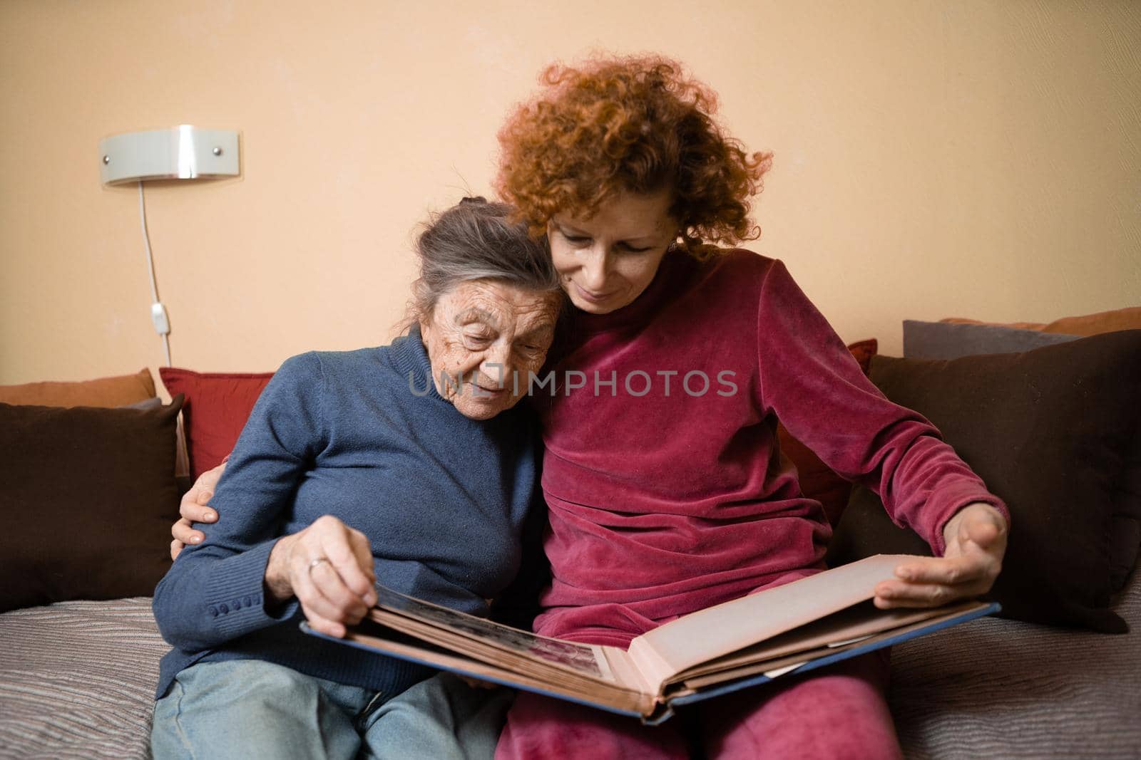 Senior woman and her adult daughter looking at photo album together on couch in living room, talking joyful discussing memories. Weekend with parents, family day, thanksgiving, mom's holiday by Tomashevska
