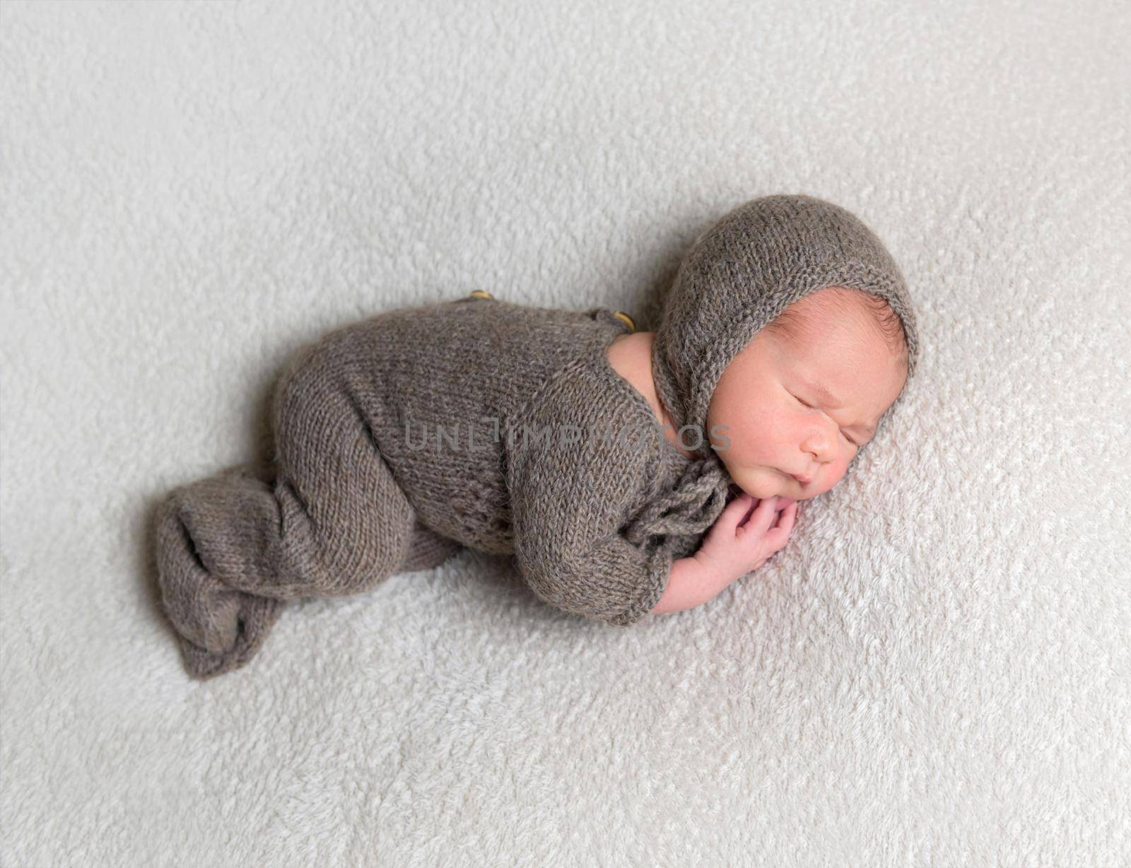 Dreamy baby boy wearing brown knitted clothes sleeps on his side on white background.