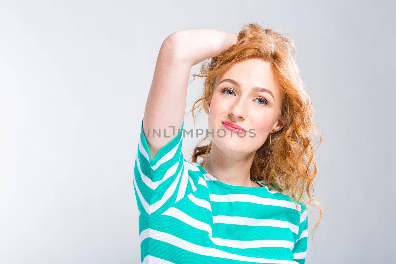 Close-up portrait of a young, beautiful woman with red curly hair in a summer dress with strips of blue in the studio on a gray background. Theme of summer vacation, tourism and summer clothes by Tomashevska