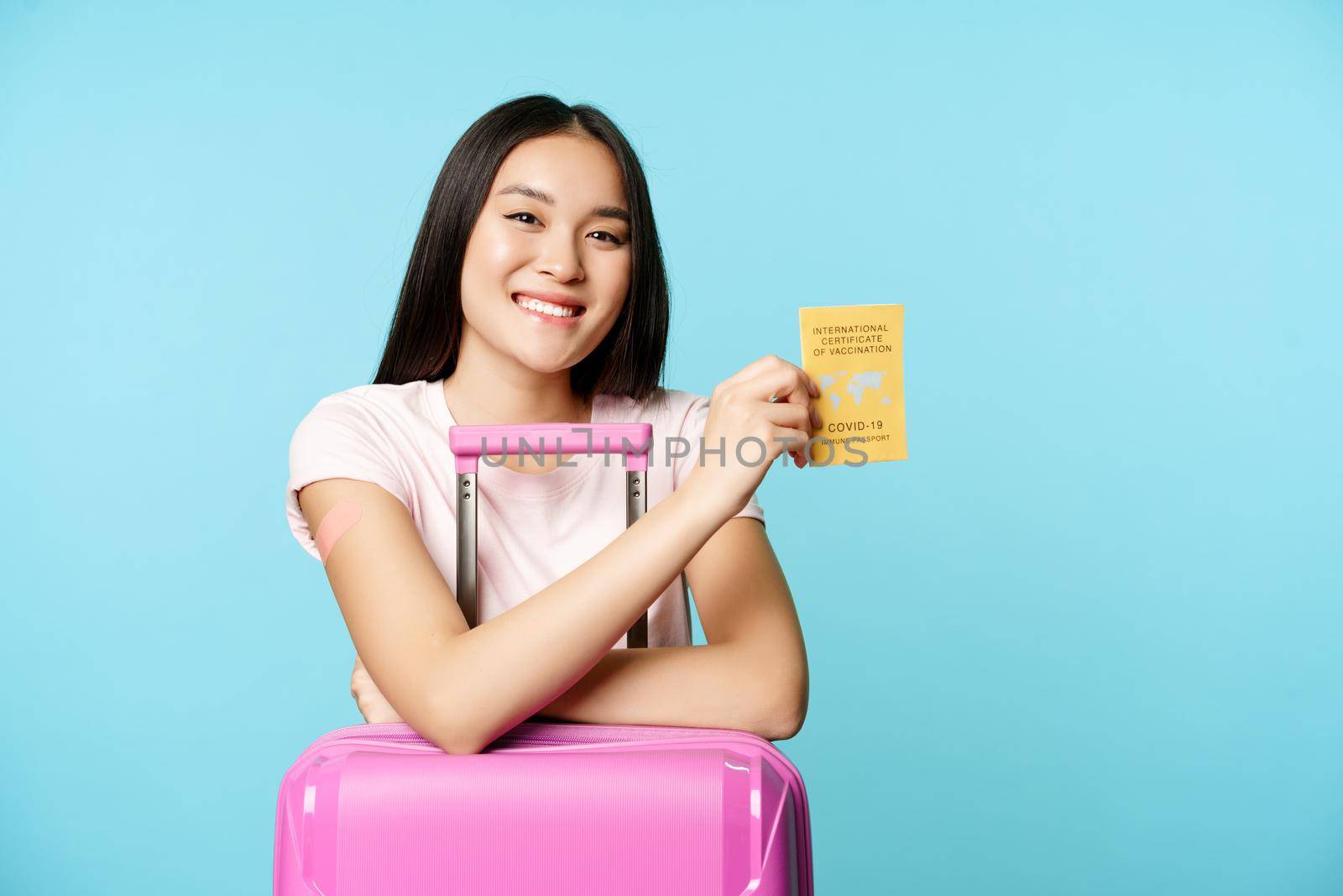 Happy smiling asian girl, tourist stands with cute suitcase in airport, shows international covid-19 vaccination certificate for travellers, travelling safe after vaccination, blue background by Benzoix