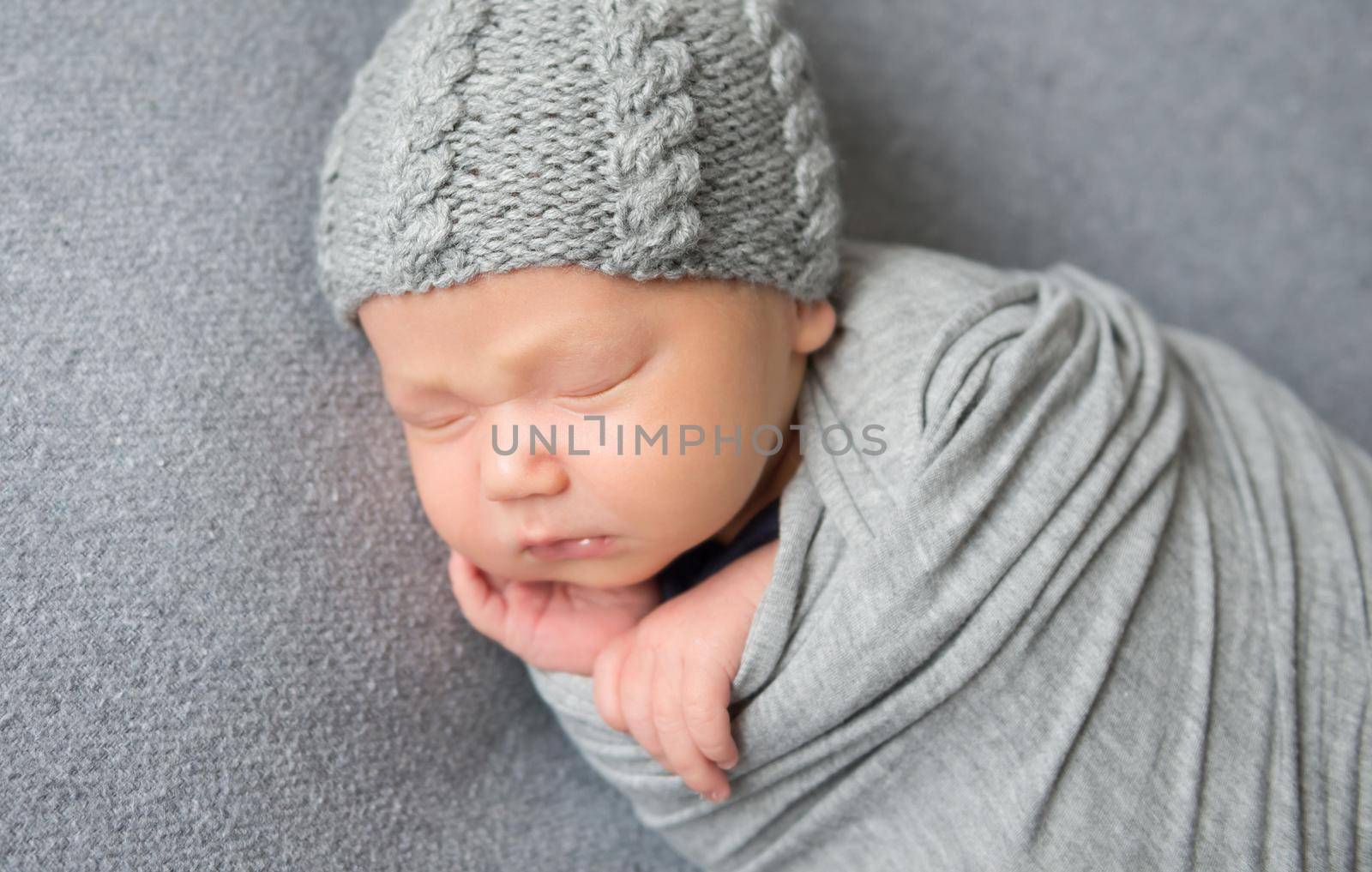 Beautiful newborn baby sleeping curled up in grey blanket and wearing grey hat. Little baby lying on grey surface