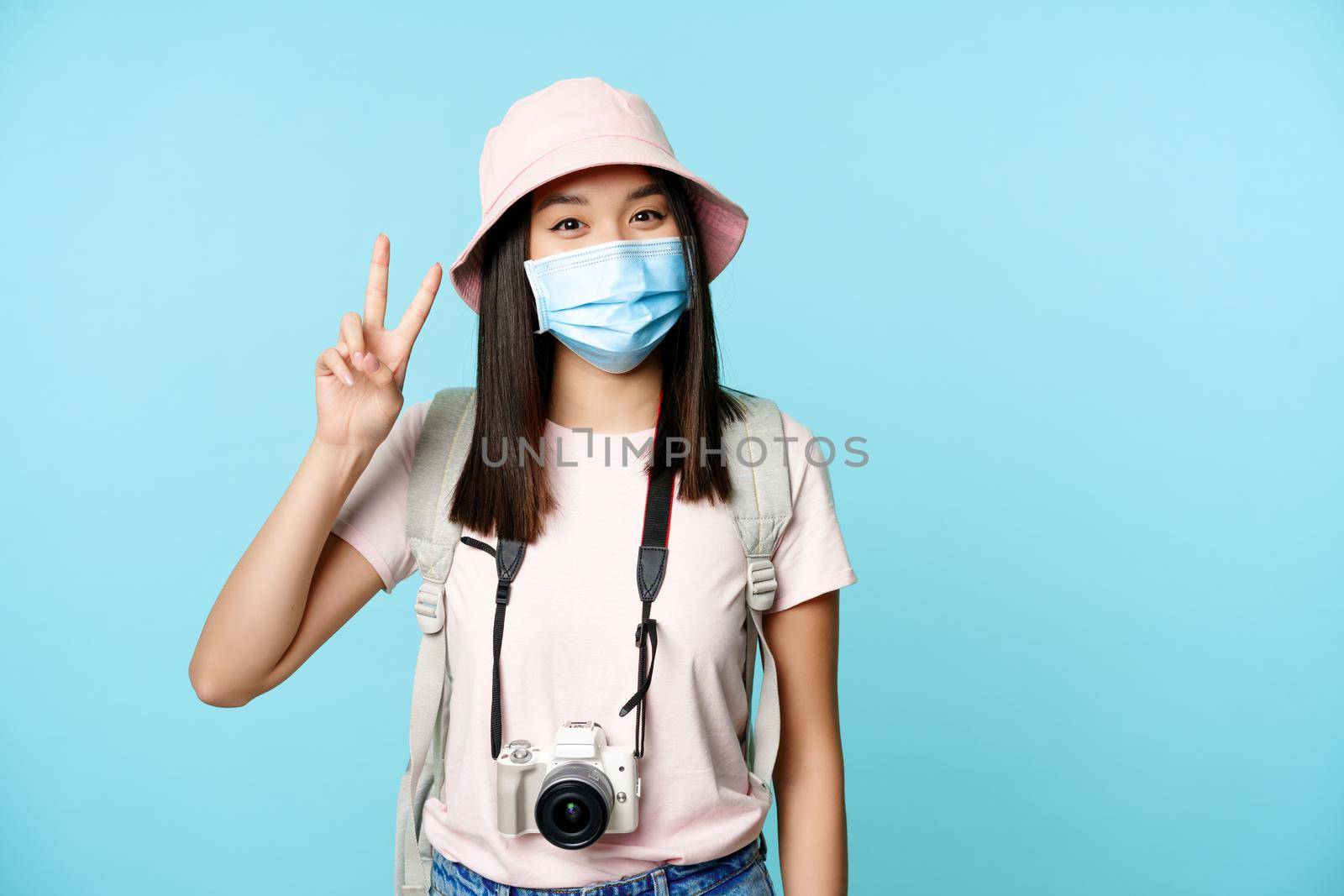 Happy korean woman in medical face mask, photo camera, showing peace v-signs, travelling during covid-19 pandemic, having vacation abroad, blue background.