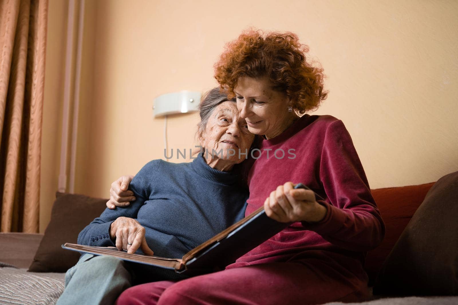 Mature daughter visits senior mother and has fun watching family photo album, sitting in embrace in living room couch. Mothers Day. Elderly woman and old gray hair grandmother laugh and remember youth