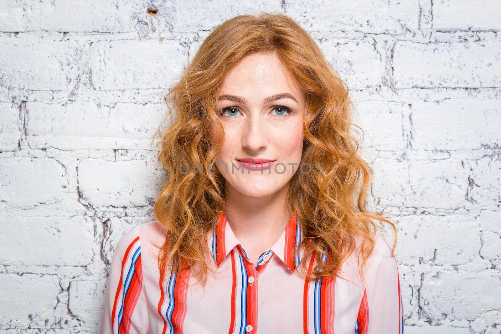 portrait of a beautiful beautiful young woman student with red curly hair and freckles on her face is leaning against a brick wall of gray color. Dressed in a red striped shirt.