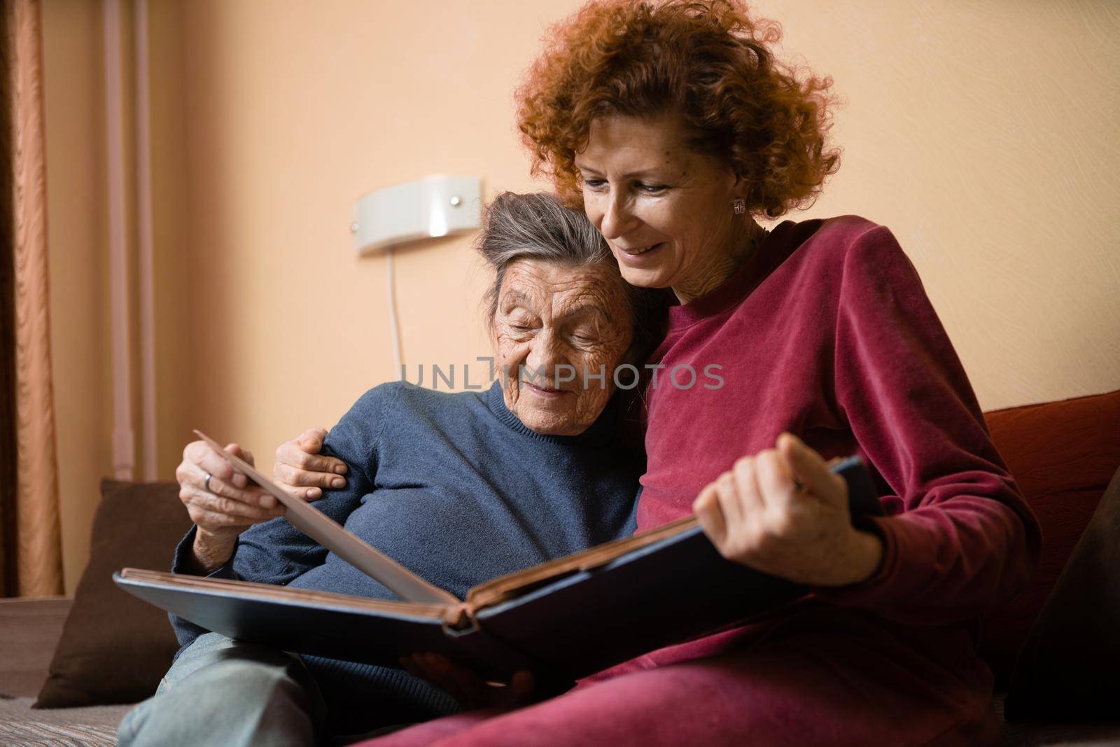 Senior woman and her adult daughter looking at photo album together on couch in living room, talking joyful discussing memories. Weekend with parents, family day, thanksgiving, mom's holiday.