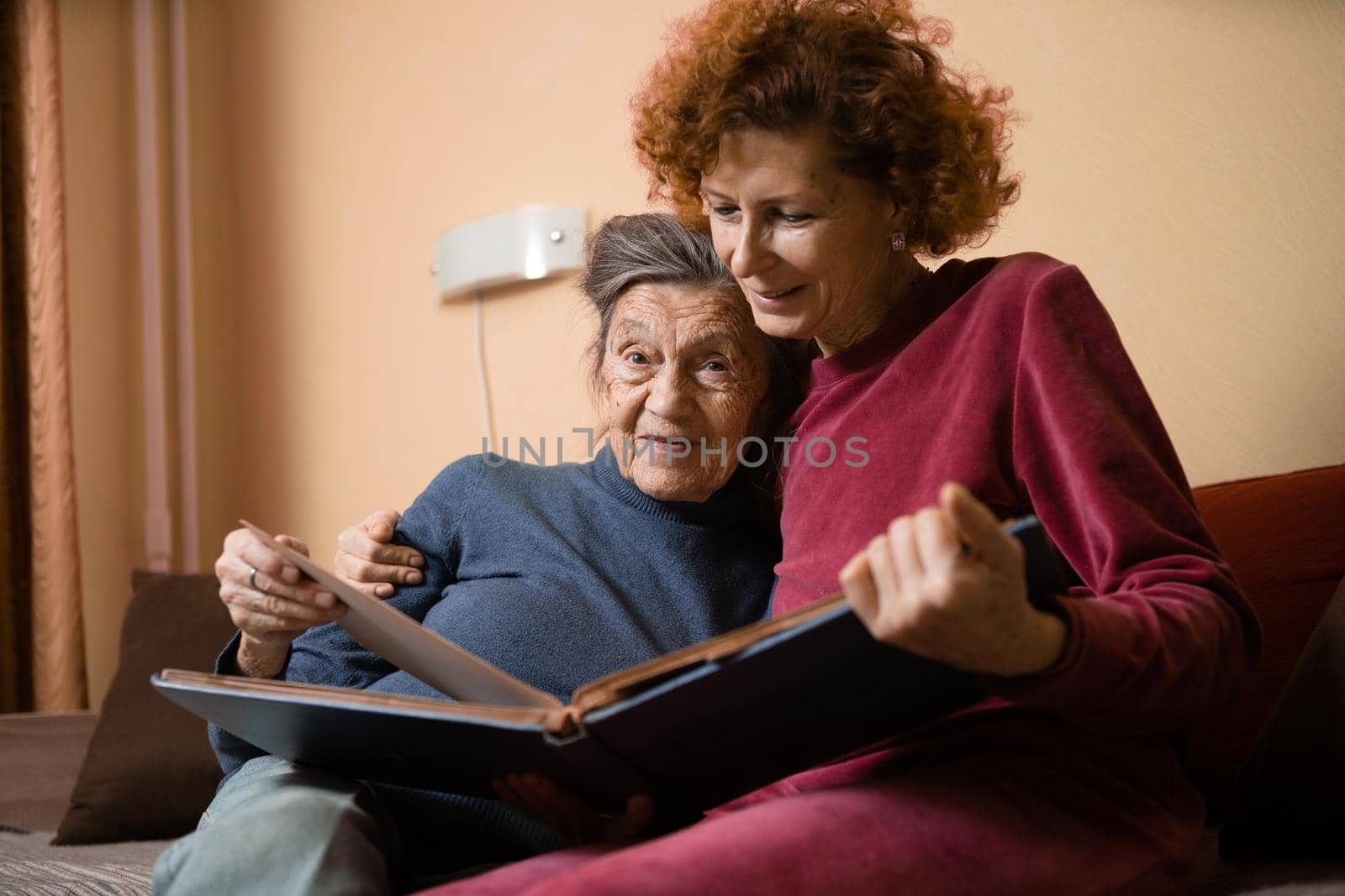 Positive aged ladies looking album photos sitting sofa at home, cheerful friends. Senior woman and her mature nurse watching photo album. Granny showing her daughter memories from the past.