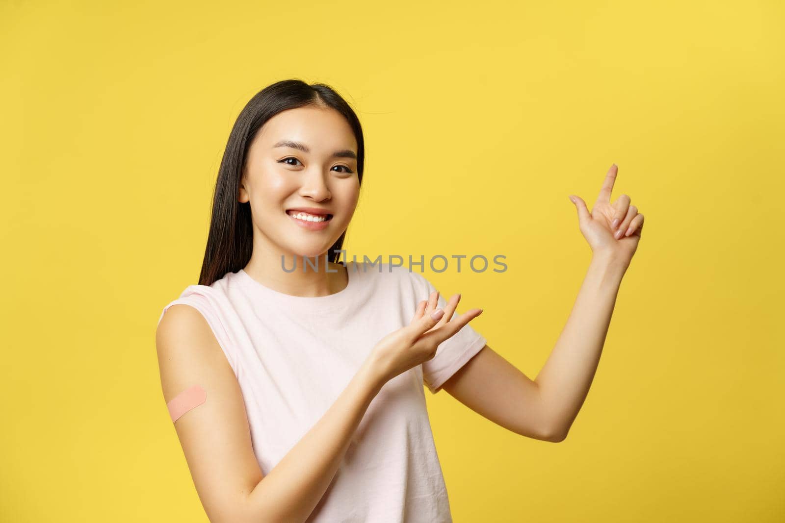 Covid-19 vaccination concept. Smiling asian vaccinated girl with patch on shoulder, pointing fingers at healthcare banner, yellow background.