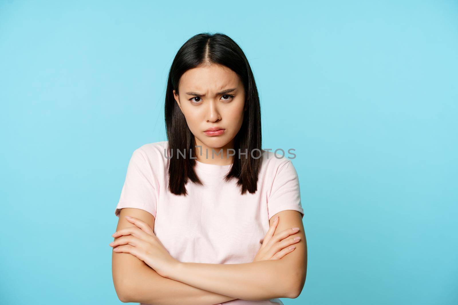 Angry teen asian girl standing in offended pose, frowning and sulking, feeling defensive, standing disappointed over blue background.