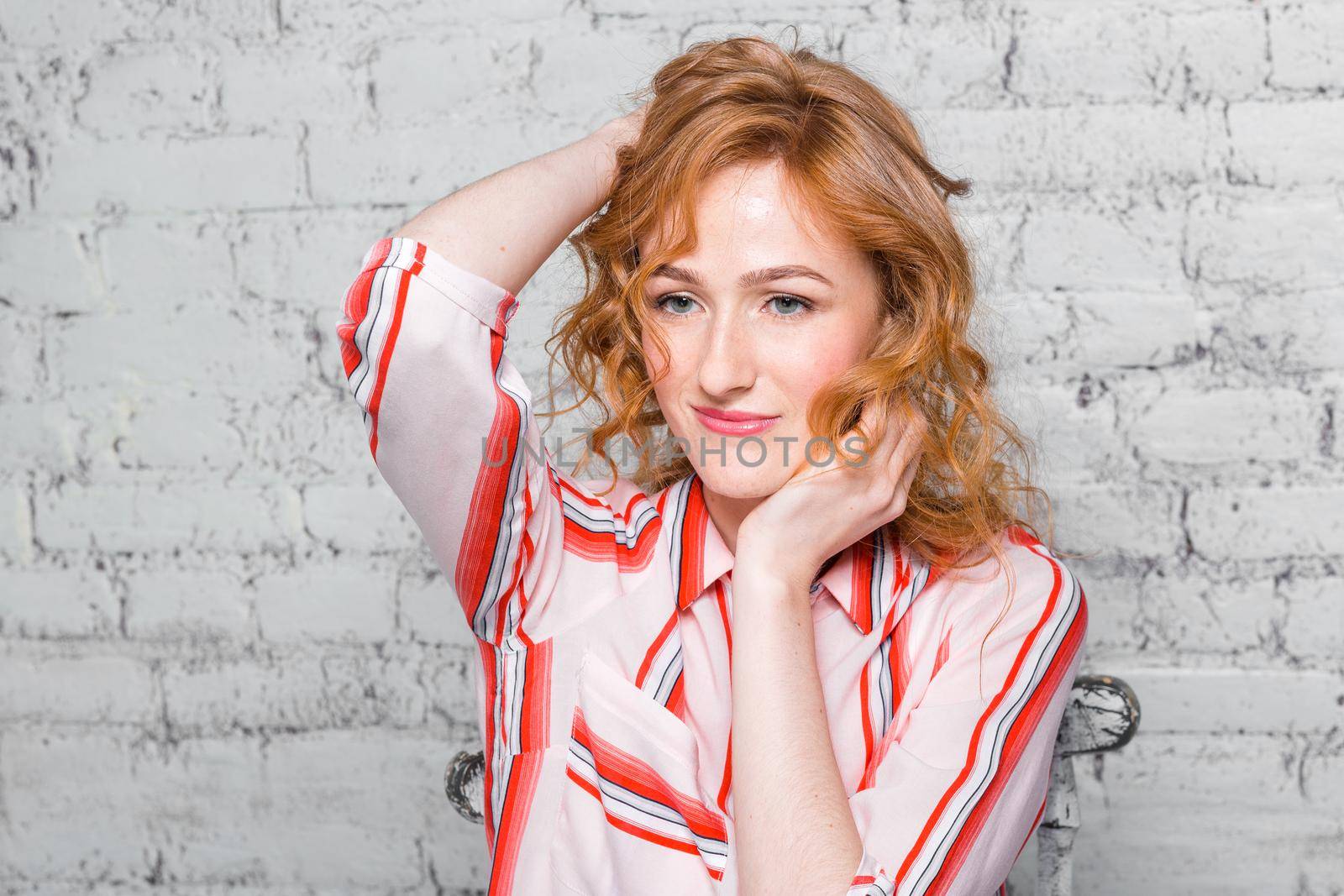 close-up portrait Beautiful young female student with red curly hair and freckles on her face sitting on a brick wall in gray background. Dressed in a red striped shirt. Hands touching the hair.