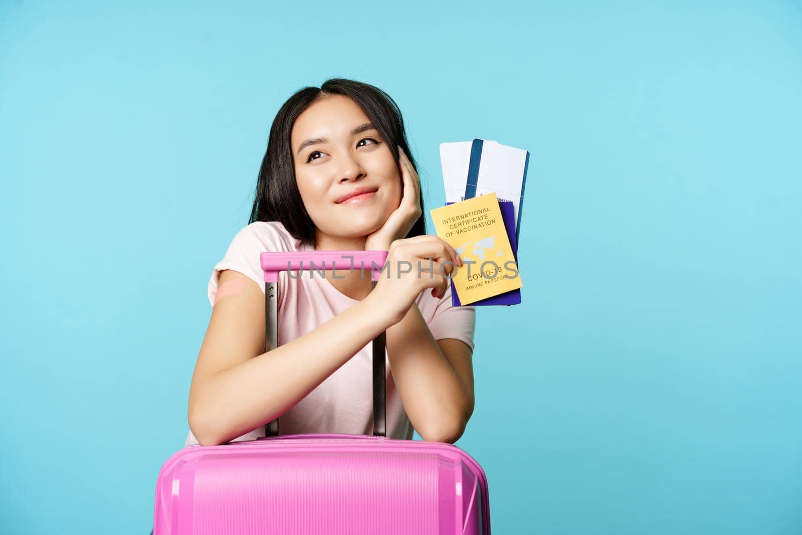 Dreamy asian tourist, smiling and thinking, holding covid-19 international vaccination certificate, two tickets and passport, standing with suitcare for trip, blue background by Benzoix