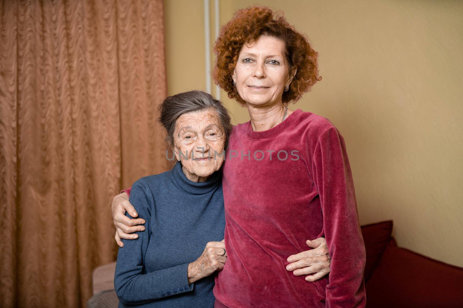 Senior Caucasian woman tenderly hugs her ninety year old elderly mother with gray hair, face with deep wrinkles, at home on sofa, smiling mother and daughter. Theme care and dedication to old parents by Tomashevska