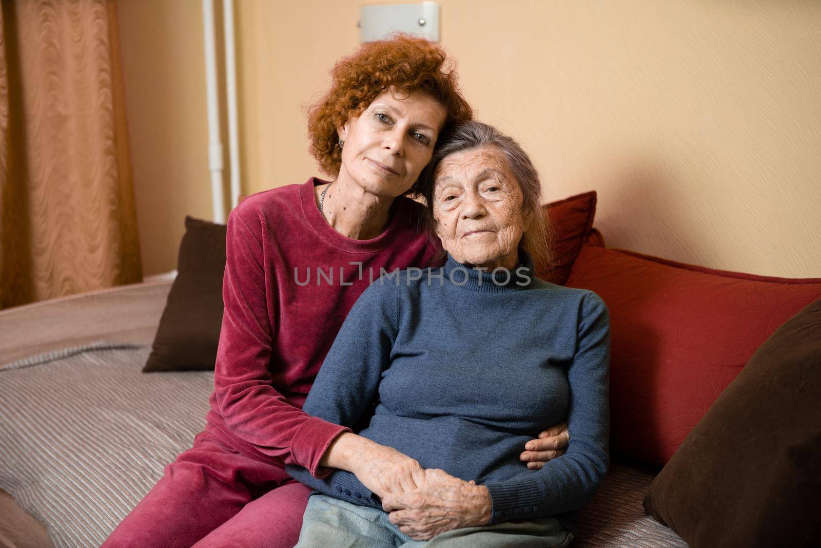 Mother and daughter, mature woman with red hair and elderly old cute happy lady woman with gray hair and deep wrinkles, embrace together at home and smile happily, family ties and caring for parents.