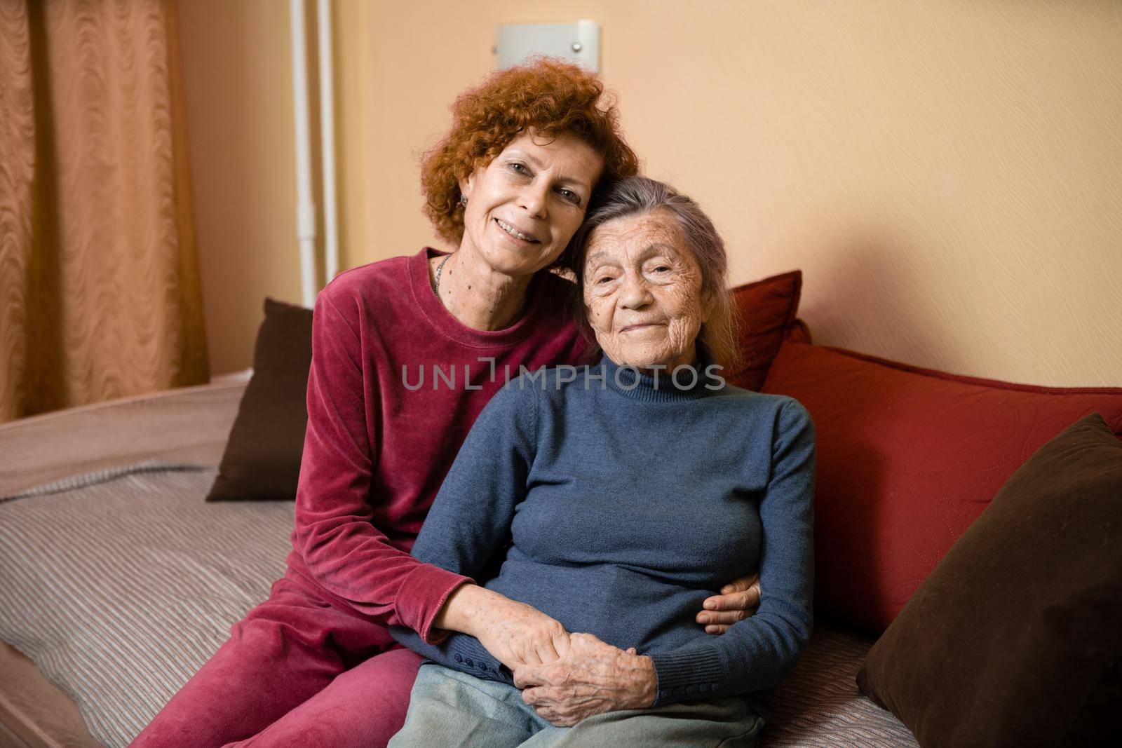 Senior Caucasian woman tenderly hugs her ninety year old elderly mother with gray hair, face with deep wrinkles, at home on sofa, smiling mother and daughter. Theme care and dedication to old parents.