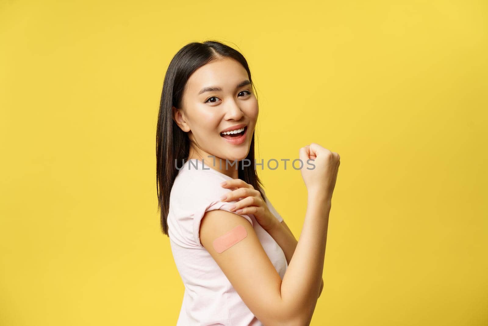 Covid-19 and healthcare medical concept. Cheerful asian girl shows arm with patch after coronavirus vaccination, getting shot of vaccine, standing happy over yellow background.
