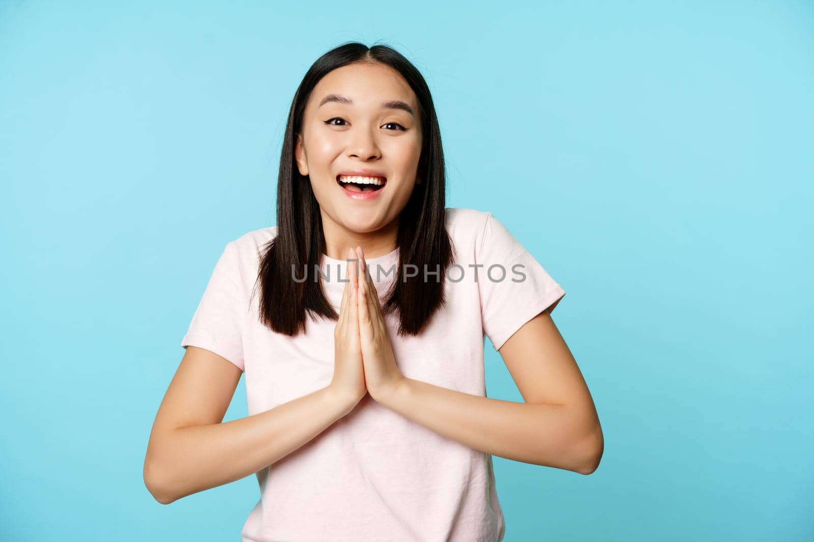 Enthusiastic asian woman looks hopeful, say please, showing begging gesture, asking for smth, standing over blue background.