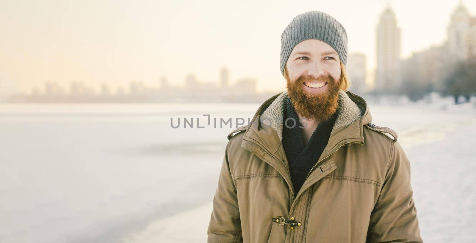 Close-up Caucasian young male red hair and beard in a hat and a park coat posing winter model against a background of a lake to freeze snow.